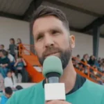 Personne tenant un micro vert, interviewée sur un terrain sportif en plein air.